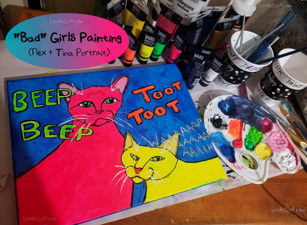 a desk littered with acrylic paint tubes and cups of water labeled "paint water don't drink" with a large painted canvas in the center featuring two dayglo kitties and the words "beep beep" "toot toot" and "waaaaahhh"